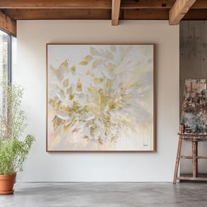 Large square abstract painting with a wooden frame. The artwork features a soft, neutral color palette of white, cream, and pale yellow, with a botanical theme. The painting is displayed in a well-lit room with a concrete floor.