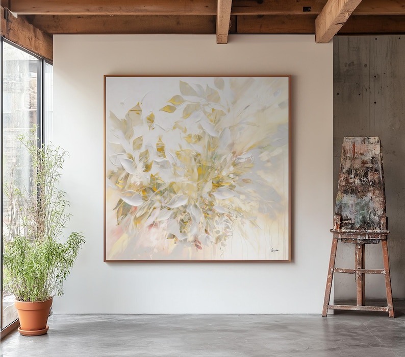 Large square abstract painting with a wooden frame. The artwork features a soft, neutral color palette of white, cream, and pale yellow, with a botanical theme. The painting is displayed in a well-lit room with a concrete floor.