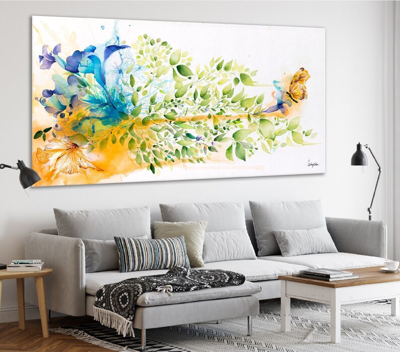 Abstract watercolor painting featuring a vibrant mix of blue, green, and yellow hues. The artwork depicts a stylized floral arrangement with a butterfly in flight. The painting is framed and displayed on a wall above a gray sofa.