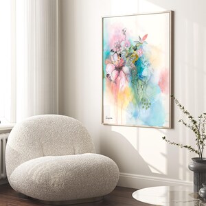 A framed watercolor painting of a bouquet of pink, blue, and green flowers. The painting is on a white background and has a light blue and yellow watercolor wash in the background. The painting is hanging on a white wall in a modern living room with a white fluffy armchair and a glass table in the foreground.