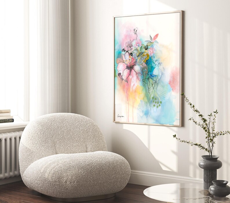A framed watercolor painting of a bouquet of pink, blue, and green flowers. The painting is on a white background and has a light blue and yellow watercolor wash in the background. The painting is hanging on a white wall in a modern living room with a white fluffy armchair and a glass table in the foreground.