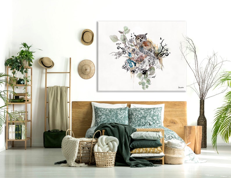 A bedroom with a wooden bed, a green patterned duvet, and a large canvas print of an owl with green leaves and branches. The canvas print is hanging on a white wall. There are two wicker baskets on the floor, a wooden ladder with a beige blanket hanging on it, and a green plant in a brown pot on the right side of the image.