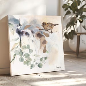 A square watercolor painting features a small brown bird perched on a branch of eucalyptus leaves. The background is a soft blend of beige, light blue, and brown watercolor washes.  The painting has a delicate, nature-inspired theme. The artist's signature is subtly placed in the bottom right corner.