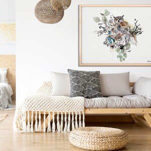 A framed watercolor painting of an owl perched on a branch with leaves and flowers. The painting is in shades of brown, green, and blue. The painting is hanging on a white wall above a light gray futon with a white throw blanket and pillows.