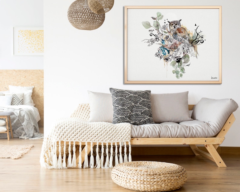A framed watercolor painting of an owl perched on a branch with leaves and flowers. The painting is in shades of brown, green, and blue. The painting is hanging on a white wall above a light gray futon with a white throw blanket and pillows.
