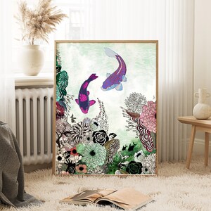 framed print of a colorful illustration with two pink and purple fish swimming in a green and blue water. The illustration is surrounded by a variety of flowers and plants in shades of green, pink, purple, and white. The print is framed in a light brown wood frame.