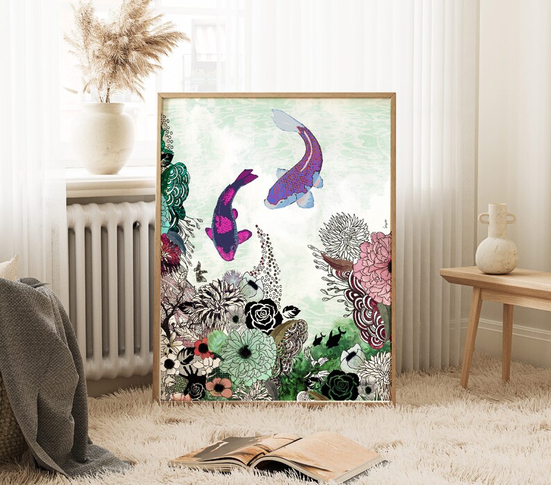 framed print of a colorful illustration with two pink and purple fish swimming in a green and blue water. The illustration is surrounded by a variety of flowers and plants in shades of green, pink, purple, and white. The print is framed in a light brown wood frame.