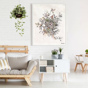 A white framed watercolor painting of a floral arrangement with birds. The painting is hanging on a white brick wall above a white cabinet with a wooden top. The cabinet has a door with a black knob and shelves with books. The cabinet is in front of a light brown futon with a white and gold striped pillow. The futon is in front of a white brick wall. There is a hanging plant with green leaves in the upper left corner of the image.