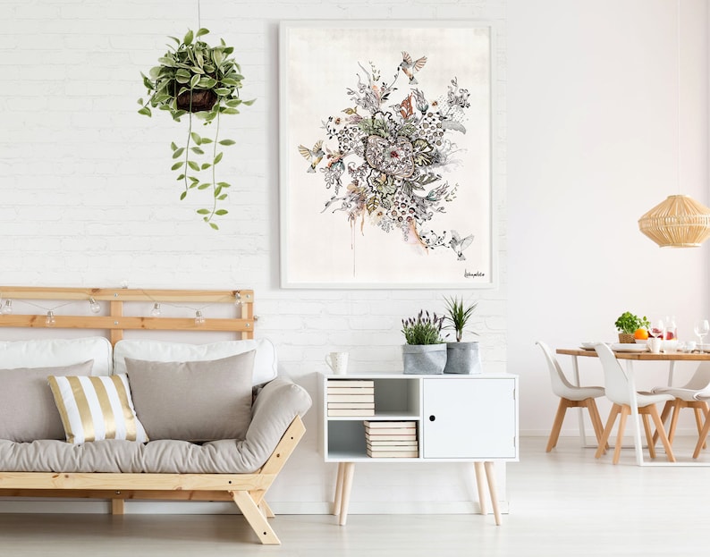 A white framed watercolor painting of a floral arrangement with birds. The painting is hanging on a white brick wall above a white cabinet with a wooden top. The cabinet has a door with a black knob and shelves with books. The cabinet is in front of a light brown futon with a white and gold striped pillow. The futon is in front of a white brick wall. There is a hanging plant with green leaves in the upper left corner of the image.
