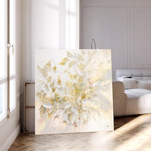 Large square canvas art featuring an abstract floral design in shades of white, cream, and gold. The artwork depicts stylized leaves and blooms, creating a soft, textured effect. The painting is displayed in a bright, modern room.