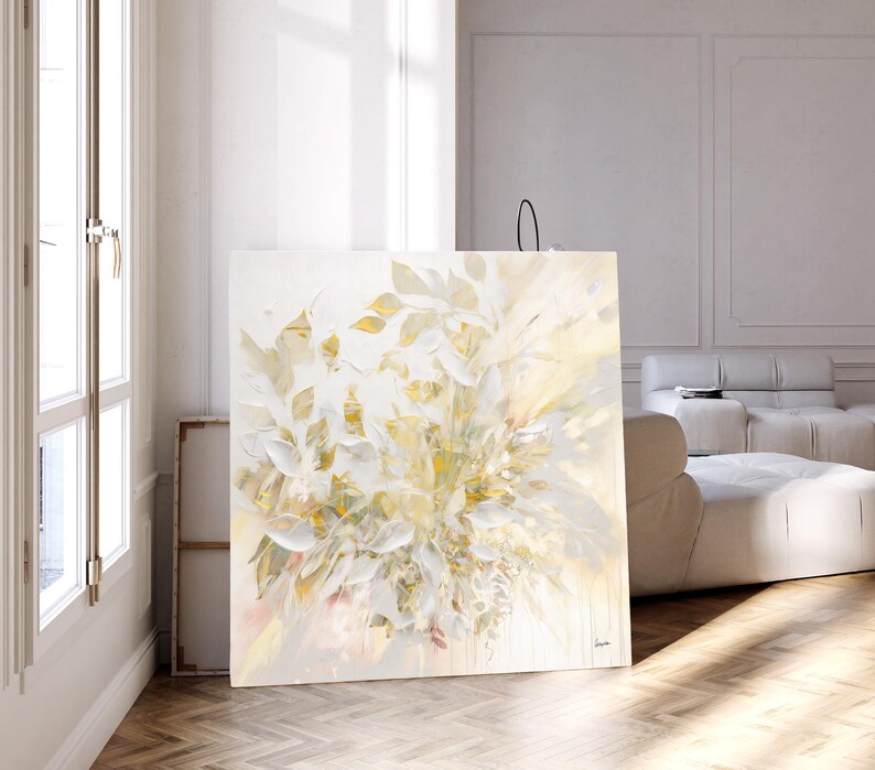 Large square canvas art featuring an abstract floral design in shades of white, cream, and gold. The artwork depicts stylized leaves and blooms, creating a soft, textured effect. The painting is displayed in a bright, modern room.