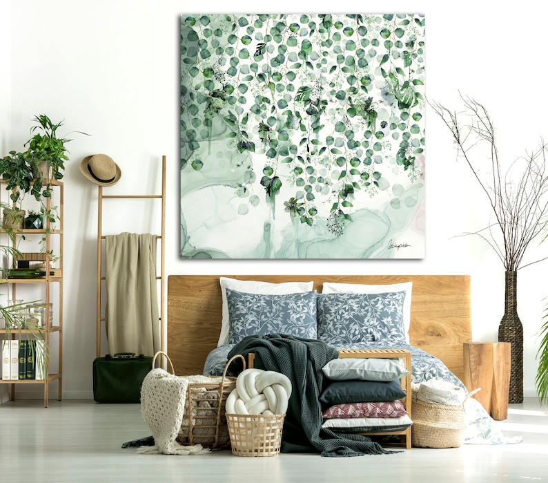 A bedroom with a wooden bed frame and a green and white floral patterned duvet. The bed has a green throw blanket and several pillows. A large painting of green leaves hangs above the bed. There are two wicker baskets and a throw blanket on the floor in front of the bed. A wooden ladder shelf with a hat and a beige throw blanket is in the background.