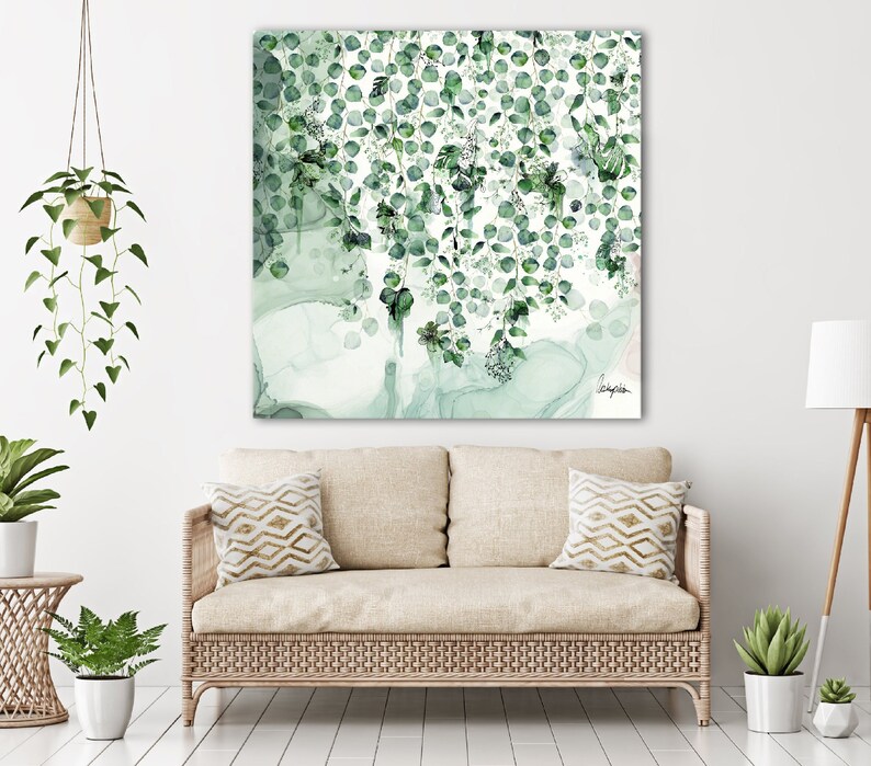 A watercolor painting of green leaves hanging from a branch. The leaves are painted in shades of green and white, and the background is a light blue watercolor wash. The painting is framed and hanging on a white wall above a beige couch.