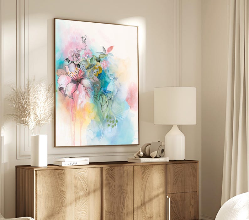 A watercolor painting of a pink flower with green leaves and blue accents on a white background. The painting is framed and hanging on a wall above a wooden dresser.
