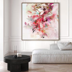 Large framed abstract floral art print. The artwork features a vibrant display of pink, red, and white flowers and leaves. The painting is set against a white background and framed in a brown wooden frame. The artwork is displayed in a modern living room setting.