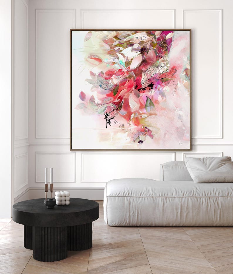 Large framed abstract floral art print. The artwork features a vibrant display of pink, red, and white flowers and leaves. The painting is set against a white background and framed in a brown wooden frame. The artwork is displayed in a modern living room setting.
