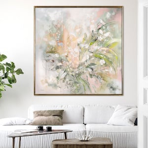 A framed abstract painting with a light pink, green, and white color scheme. The painting depicts a floral arrangement with soft, flowing brushstrokes. The painting is hanging on a white wall above a white sofa with a brown throw pillow.