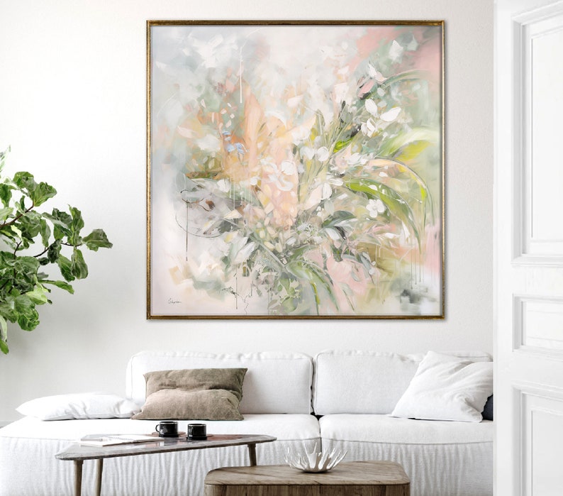 A framed abstract painting with a light pink, green, and white color scheme. The painting depicts a floral arrangement with soft, flowing brushstrokes. The painting is hanging on a white wall above a white sofa with a brown throw pillow.