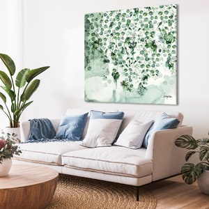 A white couch with blue throw pillows and a throw blanket. A green plant is on the left side of the couch and another green plant is on the right side. A wooden coffee table is in front of the couch. A green and white abstract painting is hanging on the wall behind the couch.