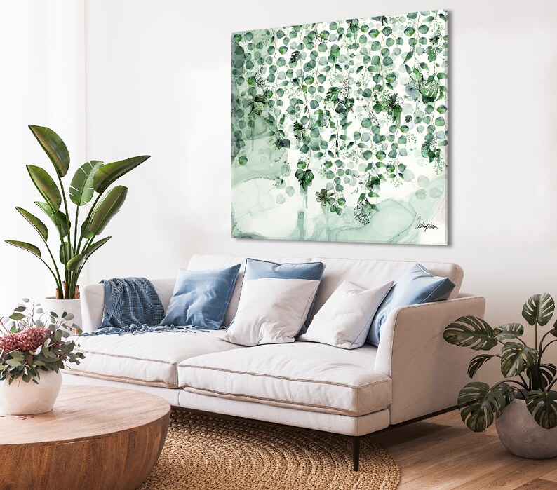 A white couch with blue throw pillows and a throw blanket. A green plant is on the left side of the couch and another green plant is on the right side. A wooden coffee table is in front of the couch. A green and white abstract painting is hanging on the wall behind the couch.