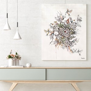A white wall with two white pendant lights hanging from the ceiling. A light wood console table with a light gray drawer sits in front of the wall. On the table are two white vases with pink flowers, a white round object, and a framed print of a black and white floral design with birds and the word 'Daphne's Botanicals' in the bottom right corner.