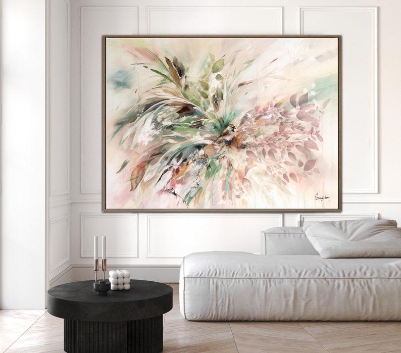 A large abstract painting with a light pink background. The painting features a large cluster of green and brown leaves in the center, with a few pink leaves scattered around the edges. The painting is framed in a light brown frame.