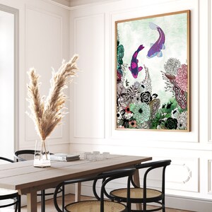 A framed illustration of two purple and pink fish swimming in a green and blue underwater scene with flowers and plants. The illustration is framed in a light wood frame and is hanging on a white wall.