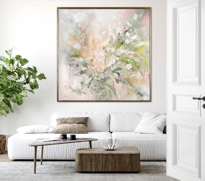A large abstract painting of white and green flowers with pink and gray accents. The painting is framed in a gold frame and is hanging on a white wall above a white couch.