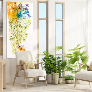A watercolor painting of a floral arrangement with blue, yellow, and green hues. The painting is framed and hanging on a white wall in a living room setting. Two white chairs with yellow pillows are placed in front of the painting, and there are plants in pots on the floor.
