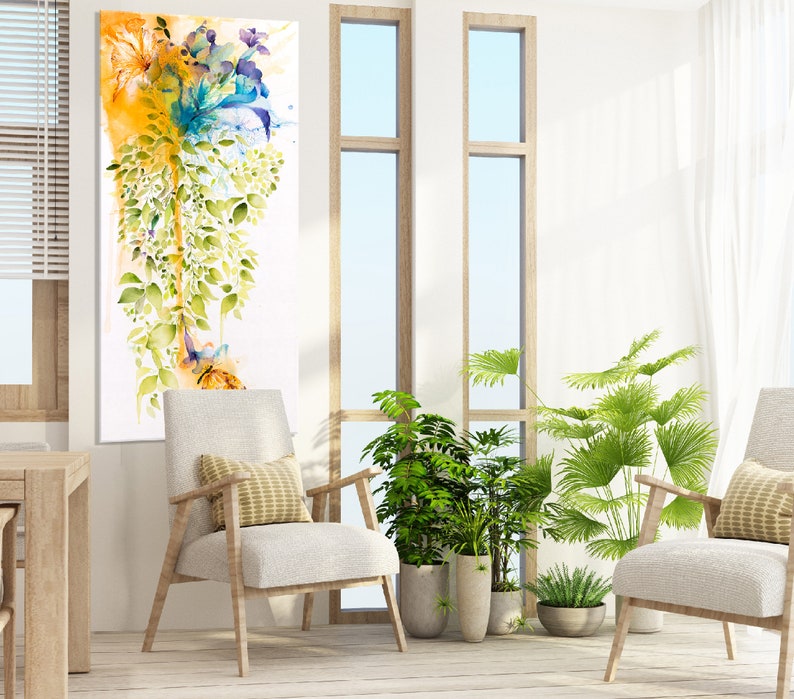 A watercolor painting of a floral arrangement with blue, yellow, and green hues. The painting is framed and hanging on a white wall in a living room setting. Two white chairs with yellow pillows are placed in front of the painting, and there are plants in pots on the floor.
