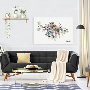 A watercolor painting of an owl perched on a branch with green leaves. The owl is brown and white with yellow eyes. The painting is framed and hanging on a white wall above a black sofa.