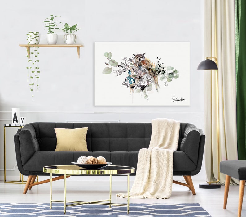 A watercolor painting of an owl perched on a branch with green leaves. The owl is brown and white with yellow eyes. The painting is framed and hanging on a white wall above a black sofa.