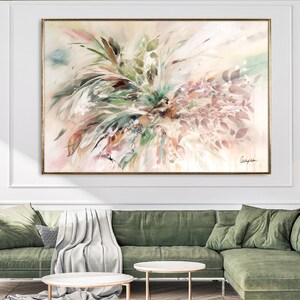 A large abstract painting with a gold frame. The painting features a mix of green, brown, and pink colors with a watercolor effect. The painting is displayed on a white wall above a green sofa.