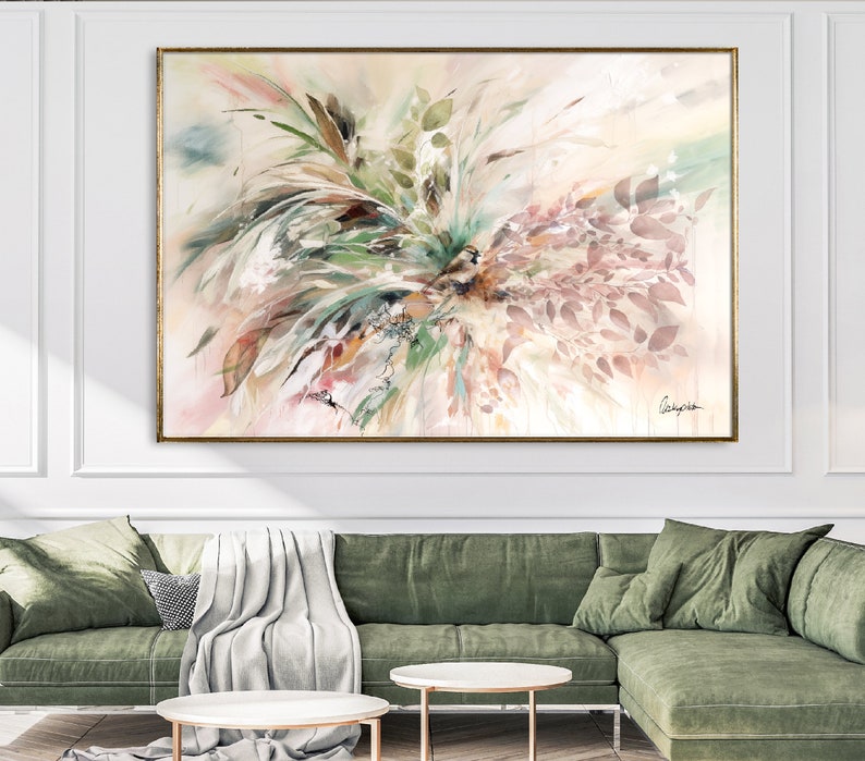A large abstract painting with a gold frame. The painting features a mix of green, brown, and pink colors with a watercolor effect. The painting is displayed on a white wall above a green sofa.