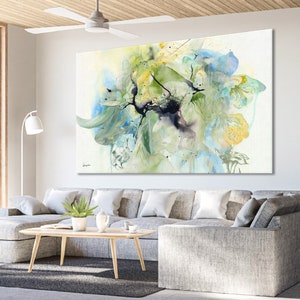 Abstract watercolor painting featuring a large, green, blue, and yellow floral design on a white background. The painting is displayed on a wall in a living room setting.