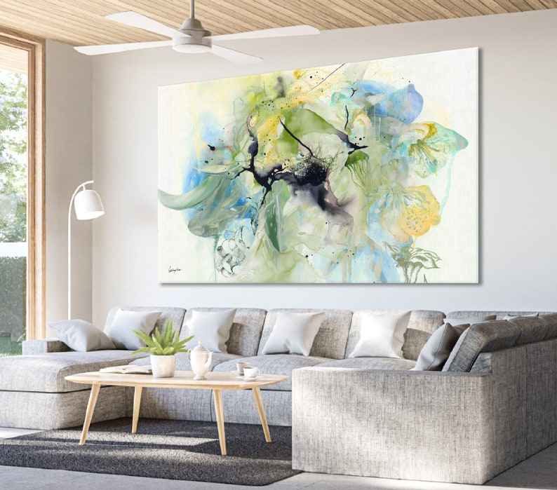 Abstract watercolor painting featuring a large, green, blue, and yellow floral design on a white background. The painting is displayed on a wall in a living room setting.