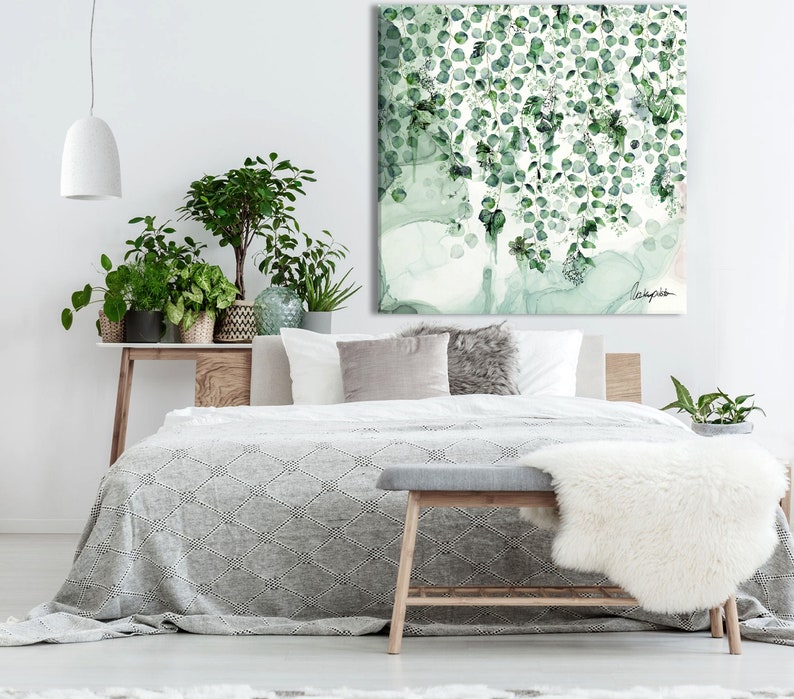 A bedroom with a white bed with a gray blanket and a wooden bench with a white faux fur throw. The bed is against a white wall with a large green and white abstract painting hanging above the bed. There are several potted plants in the room, including a large green plant on a wooden table next to the bed.