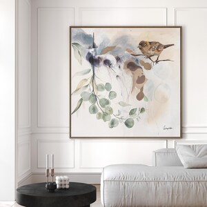 A square watercolor painting features a bird perched on a branch with eucalyptus leaves. The artwork is done in soft, muted tones of beige, brown, and green, creating a serene and calming atmosphere. The painting's style is abstract, with loose brushstrokes and flowing lines. The overall theme is nature-inspired, with a focus on organic shapes and textures.