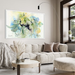 A white framed abstract watercolor painting with shades of blue, green, and yellow. The painting is hanging on a white wall above a white sofa with pillows and a throw blanket.