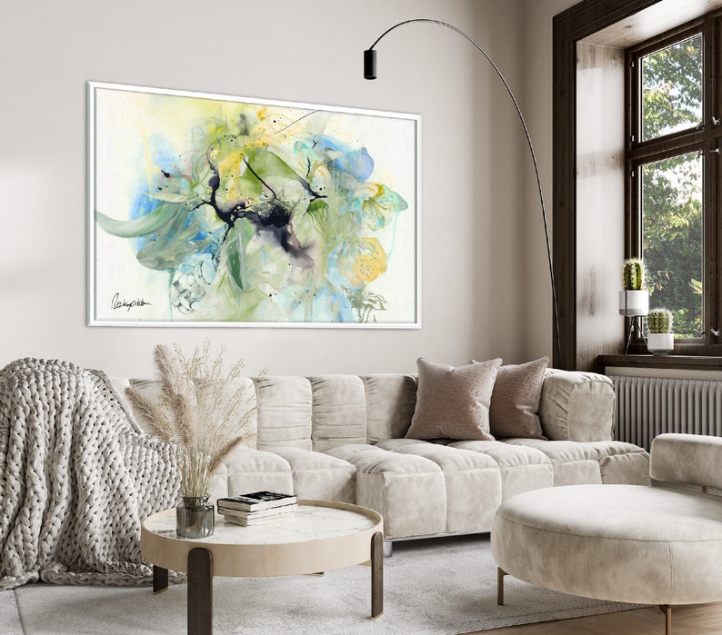A white framed abstract watercolor painting with shades of blue, green, and yellow. The painting is hanging on a white wall above a white sofa with pillows and a throw blanket.