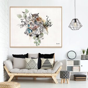 A framed watercolor painting of an owl perched on a branch with leaves and flowers. The painting is in a light wood frame and is hanging on a white wall above a light brown couch with black and white patterned pillows. There is a black metal cage light fixture hanging from the ceiling above the couch. There is a small white cabinet with a black and white patterned box on the bottom shelf.