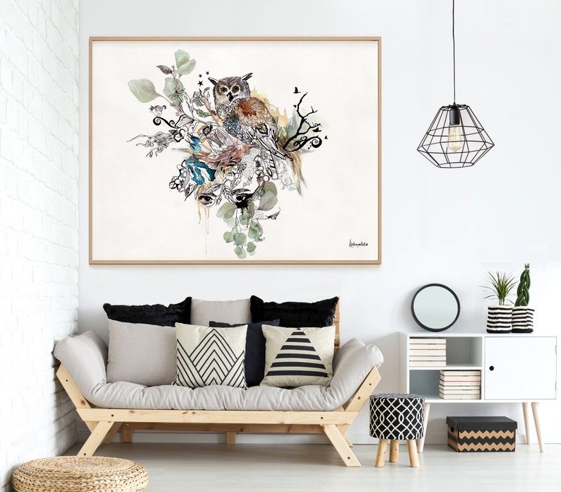 A framed watercolor painting of an owl perched on a branch with leaves and flowers. The painting is in a light wood frame and is hanging on a white wall above a light brown couch with black and white patterned pillows. There is a black metal cage light fixture hanging from the ceiling above the couch. There is a small white cabinet with a black and white patterned box on the bottom shelf.