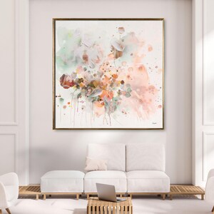 Large framed abstract watercolor painting with a gold frame. The artwork features a soft color palette of pink, peach, green, and brown hues. The painting is displayed above a white sofa and ottoman.