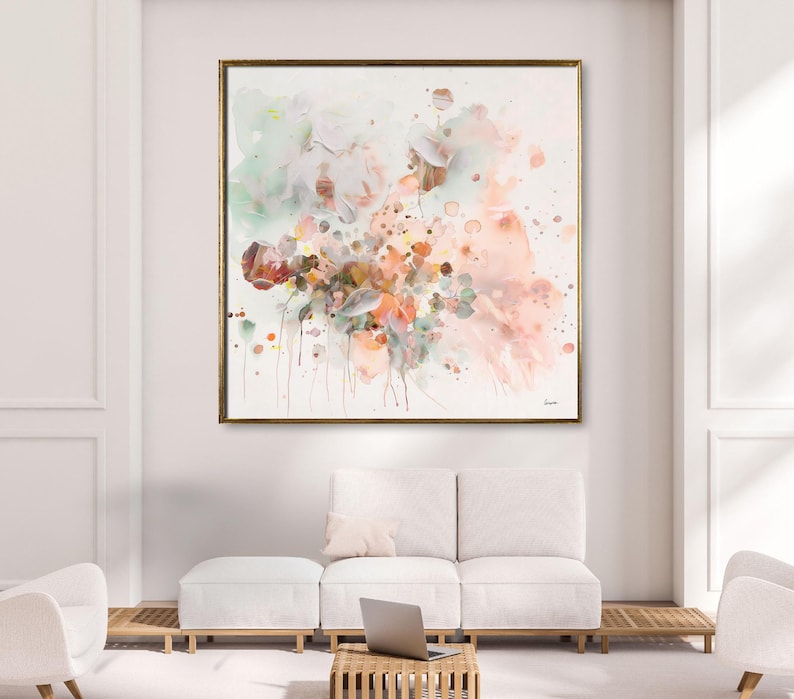 Large framed abstract watercolor painting with a gold frame. The artwork features a soft color palette of pink, peach, green, and brown hues. The painting is displayed above a white sofa and ottoman.