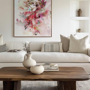 A living room scene featuring a large abstract floral painting in a wooden frame above a white sofa. A wooden coffee table holds two white vases and books. Decorative pillows and neutral tones create a cozy atmosphere.