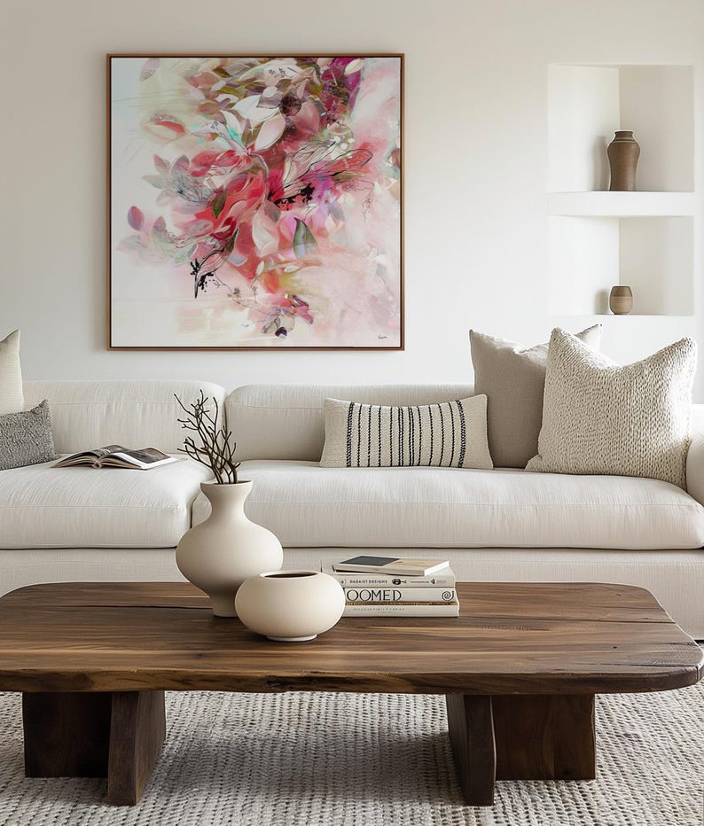 A living room scene featuring a large abstract floral painting in a wooden frame above a white sofa. A wooden coffee table holds two white vases and books. Decorative pillows and neutral tones create a cozy atmosphere.