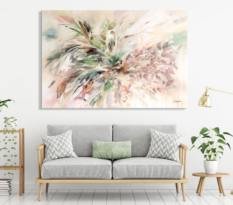 Abstract watercolor painting with a light pink, beige, and green color scheme. The painting features a cluster of leaves and branches in the center of the canvas. The leaves are painted in shades of green, brown, and pink. The branches are painted in shades of brown and beige. The painting is displayed on a wall above a gray sofa with green and white pillows.