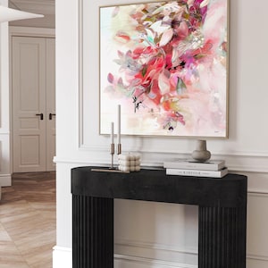 An abstract floral art print with a gold frame. The artwork features a blend of pink, red, and white hues, with black accents. The print hangs above a black console table with fluted detailing. On the table are candles, a decorative cube, and books.