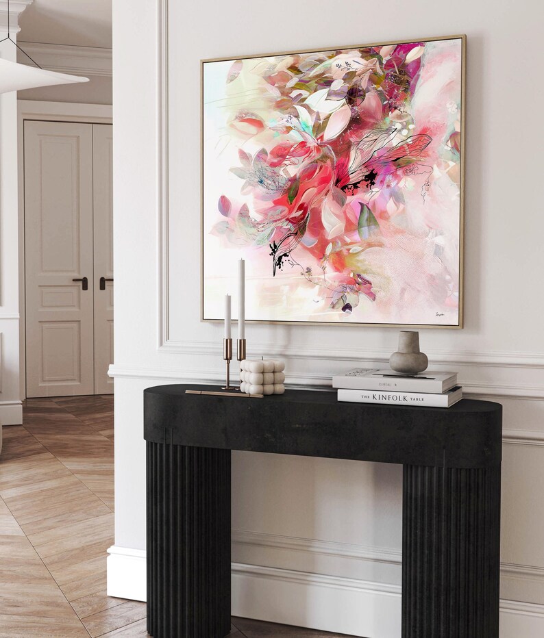An abstract floral art print with a gold frame. The artwork features a blend of pink, red, and white hues, with black accents. The print hangs above a black console table with fluted detailing. On the table are candles, a decorative cube, and books.
