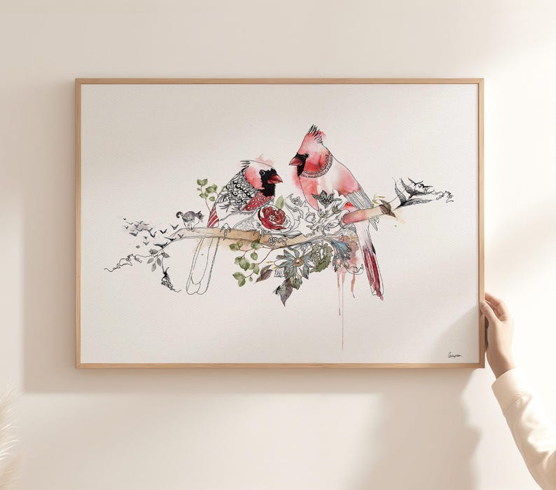 A framed horizontal art print featuring two red cardinal birds perched on a branch. The illustration combines watercolor washes in shades of red and pink with intricate black ink line work, depicting floral elements and foliage.